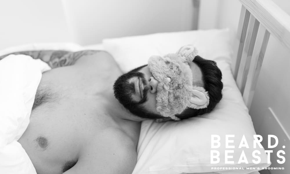 Man with a full beard sleeping on a white pillow while wearing a fluffy sleep mask, representing overnight beard care