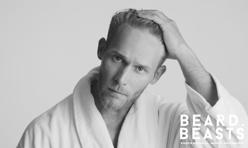 Man in a white bathrobe gently styling damp hair, illustrating the importance of proper moisture levels before applying hair product.
