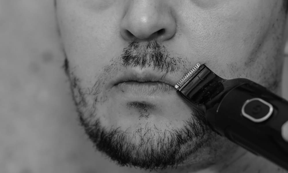 Close-up of a man trimming his mustache unevenly with an electric trimmer, showing a common grooming mistake around the lip line.