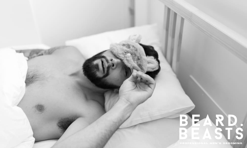 Shirtless man lying in bed with a sleep mask, showing relaxed sleep posture—ideal for illustrating beard protection during sleep.