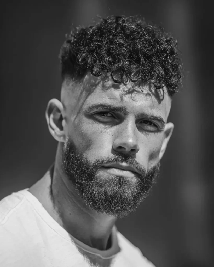 Short curly top with a skin fade and beard, showing defined curls above tight faded sides with fuller facial hair.