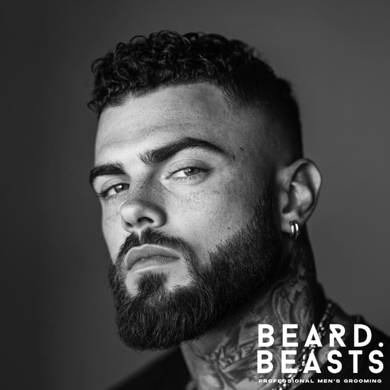 Black and white image of a man with a short curly top and skin fade, paired with a full beard—one of the boldest short haircuts for men with beards.