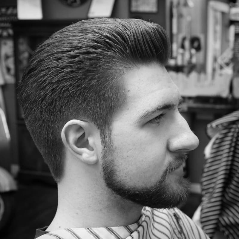 Side profile of a man with a short pompadour hairstyle and well-groomed beard in a barbershop setting.