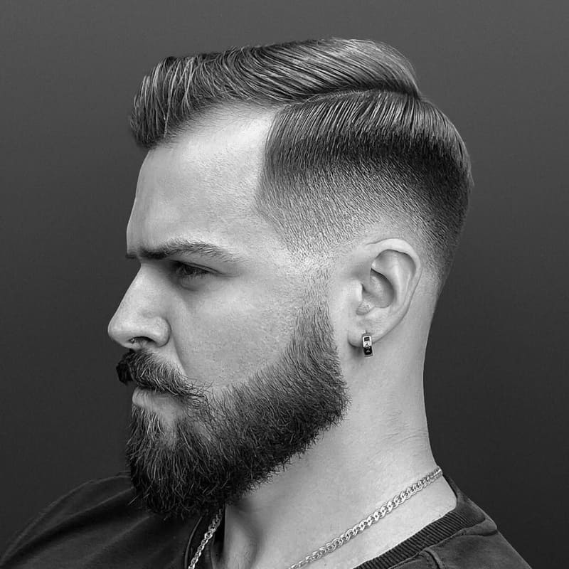 Black and white portrait of a man with a short side part haircut and beard, featuring a clean fade and defined part—classic style for men’s grooming.