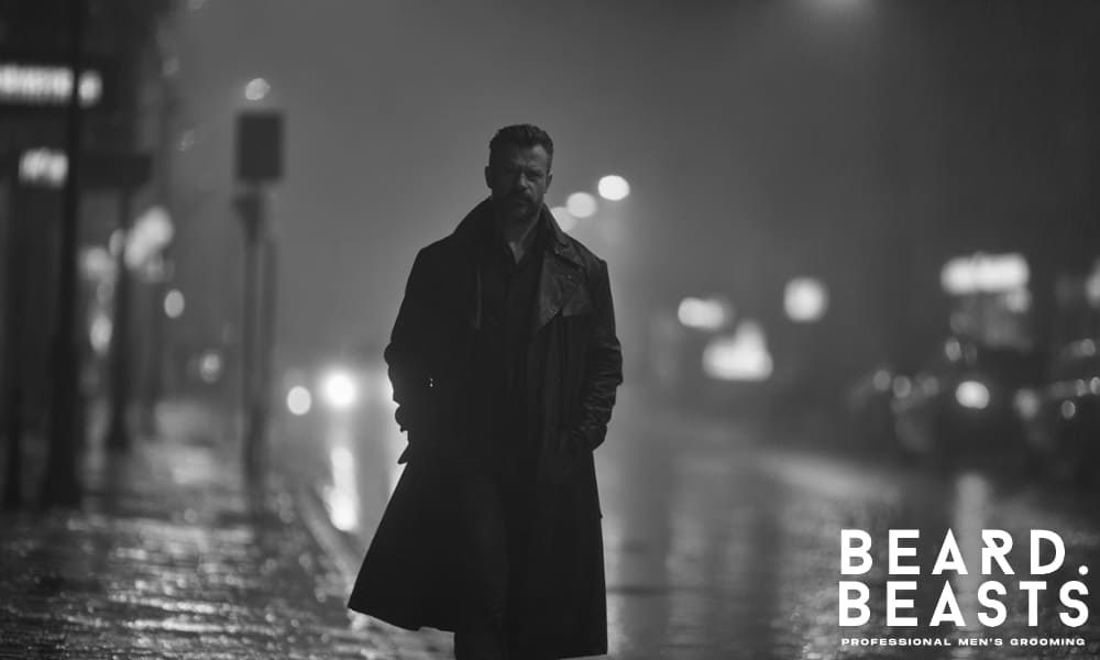 A serious bearded man walking alone at night in a dark trench coat on a wet city street, under moody streetlights.