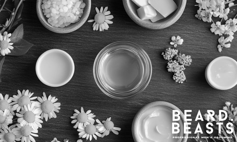 Flat lay of natural beard wash ingredients including chamomile flowers, shea butter, aloe vera, and essential oils on a dark slate background, showcasing the key components that make beard wash effective.