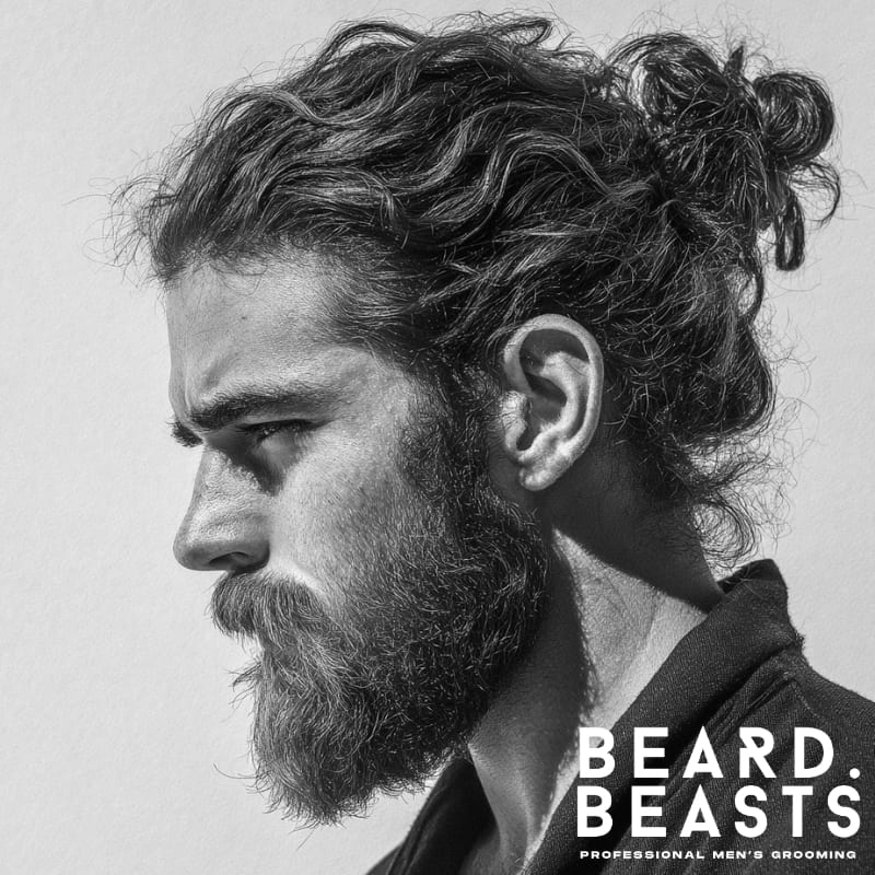 Black and white side portrait of a rugged man with a long, textured man bun and a thick full beard, featuring natural waves and clean grooming—bold, confident style with refined structure.