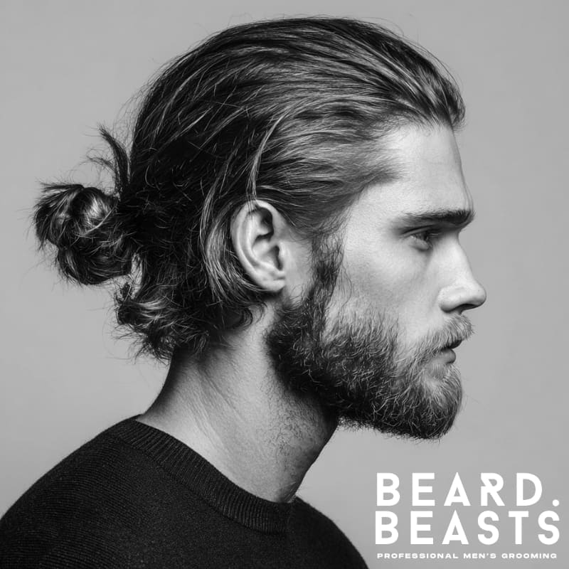 Side profile of a man with a low man bun tied at the nape of the neck and a full, well-groomed beard, featuring sleek, combed-back hair and a clean neckline—refined and minimalist grooming style.