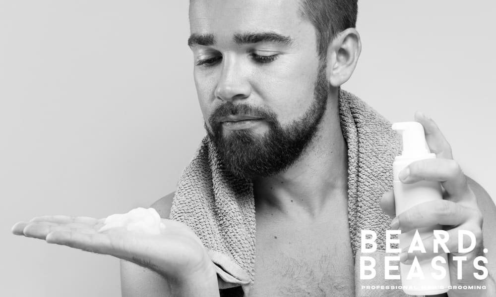 Bearded man with a towel over his shoulders dispensing foaming beard wash into his hand, highlighting a daily grooming routine for clean, healthy facial hair.