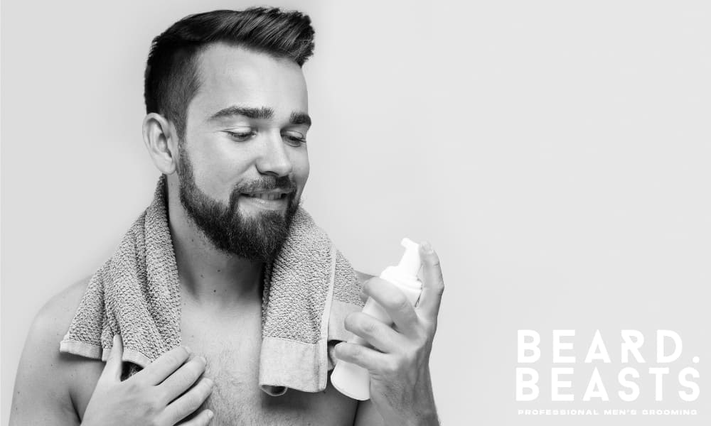 Smiling bearded man with a towel around his neck holding a foaming beard wash bottle, representing a clean and confident grooming routine with copy space on the left.