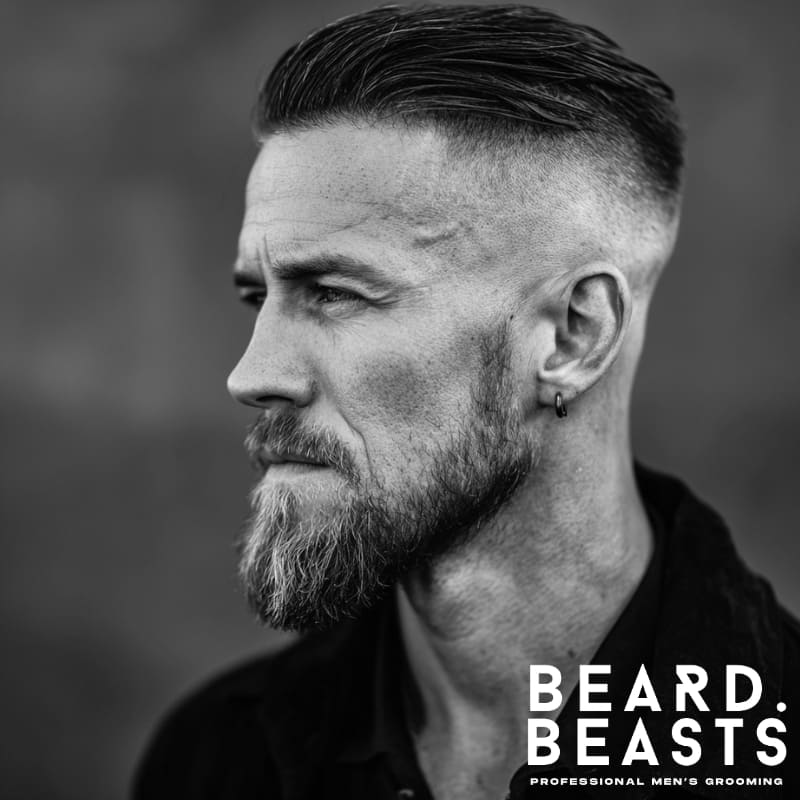 Man with a slicked-back undercut and short boxed beard, wearing an earring—classic masculine grooming style in black and white.