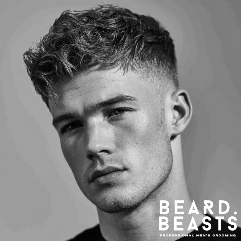 Professional young male model with a high fade wavy textured crop, featuring defined waves on top and a bold high fade on the sides. The hair on top is styled with soft, tousled waves that add volume and texture, while the sides are sharply faded, creating a clean contrast. The overall look is modern and edgy, with the high fade highlighting the full texture of the top.