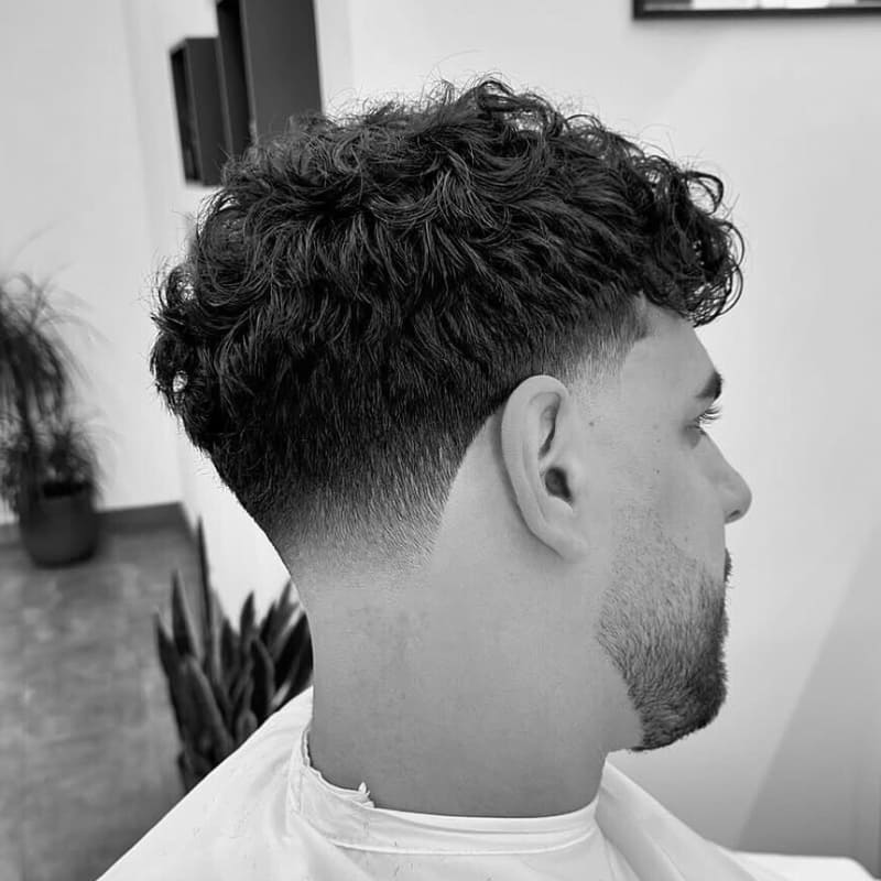 Professional young male model with a wavy textured crop and taper fade. The hair on top is styled with natural, defined waves, while the sides gradually fade from short to shorter, creating a smooth, seamless transition. The tapered sides add a sharp contrast to the voluminous, textured top, resulting in a modern and refined look.