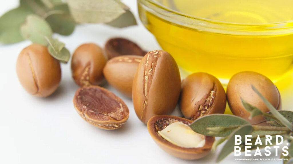 Close-up of argan seeds with a glass bowl of golden argan oil, a nutrient-rich carrier oil for beard care and grooming.