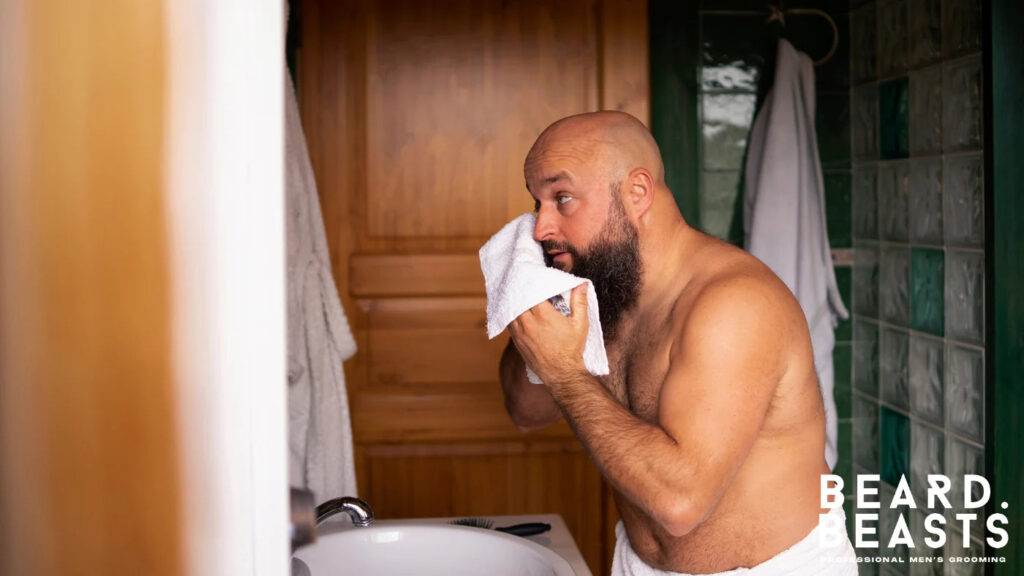 Bald man with a full beard drying his face and beard with a towel in front of the mirror after washing, part of a beard grooming routine.