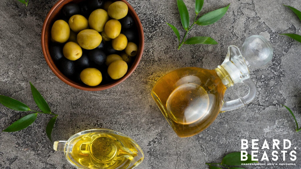 Glass jug of olive oil with a bowl of black and green olives on a stone background, a natural carrier oil for beard hydration and protection.