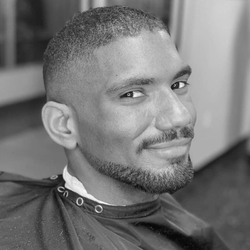 A black and white portrait of a man with an oblong face shape, featuring a well-groomed chin strap beard with a mustache. The beard is precisely trimmed along the jawline, creating a sharp and structured look that enhances facial definition without adding unnecessary length. The mustache is neatly shaped, complementing the chin strap to add balance to the face. His short, faded haircut further accentuates the clean and polished appearance. The image is taken in a barbershop setting, highlighting a professional grooming style ideal for oblong faces.
