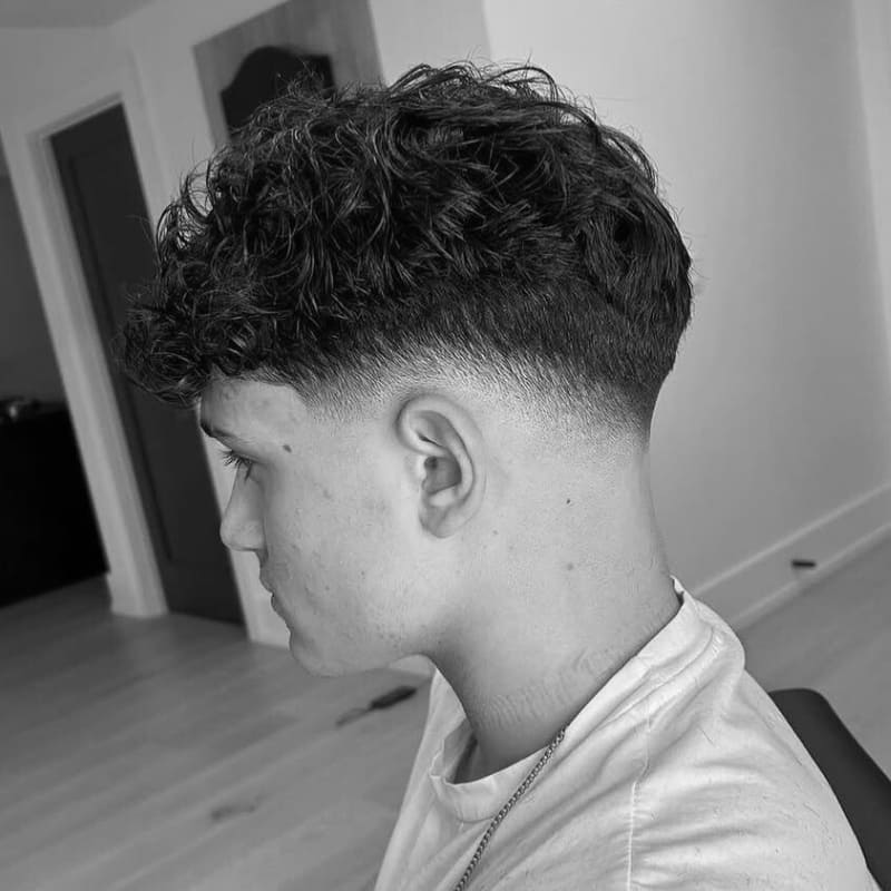 A black-and-white image of a man with a curly hairstyle. The top of his hair is styled with soft, defined curls, while the sides are blended neatly, tapering to a close buzz around the ears.