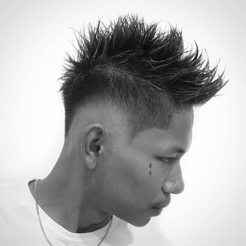 Man with a sharp drop fade faux hawk haircut featuring spiked textured hair on top and a curved fade that dips behind the ear for an edgy, modern look.