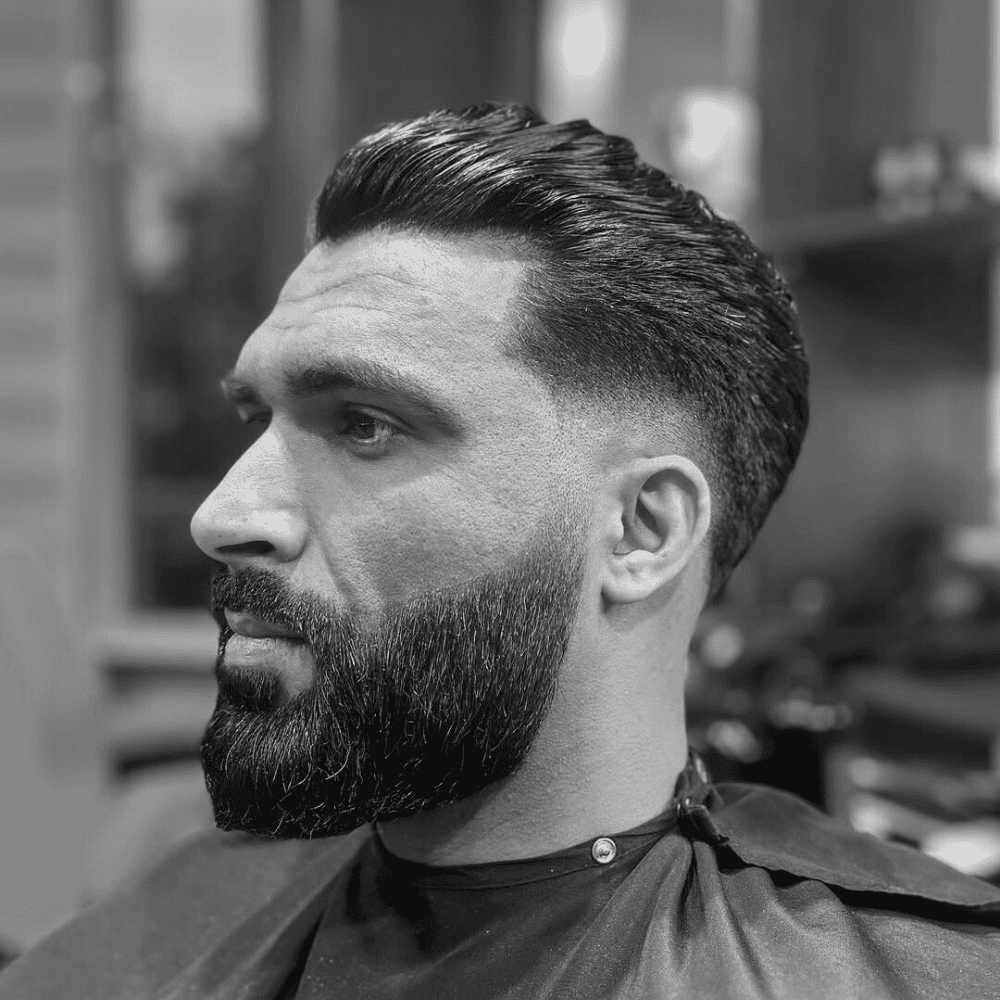 A three-quarter profile shot of a man in a barber's chair, showcasing a slick back haircut with a low fade. The voluminous hair on top is combed back, and the sides have a low, clean taper fade that blends into his full beard.