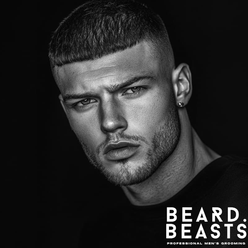 A young man is shown with a micro fringe haircut, characterized by very short, straight-across bangs that sit high on his forehead. His hair is textured and slightly wavy, styled close to the scalp with a natural, tousled look that adds a casual, effortless vibe to the overall appearance. The micro fringe creates a bold and minimalistic statement, contrasting with the slightly longer hair on the sides and back. He is wearing dark sunglasses with a thin, gold-accented frame, and his expression is relaxed and contemplative. Dressed in a green shirt, the man appears to be indoors, near a large window that lets in soft, natural light, which highlights the texture and detail of his hair. The clean lines of the micro fringe, paired with the casual waves and sunglasses, give him a modern and stylish look, ideal for someone seeking a unique and low-maintenance haircut.