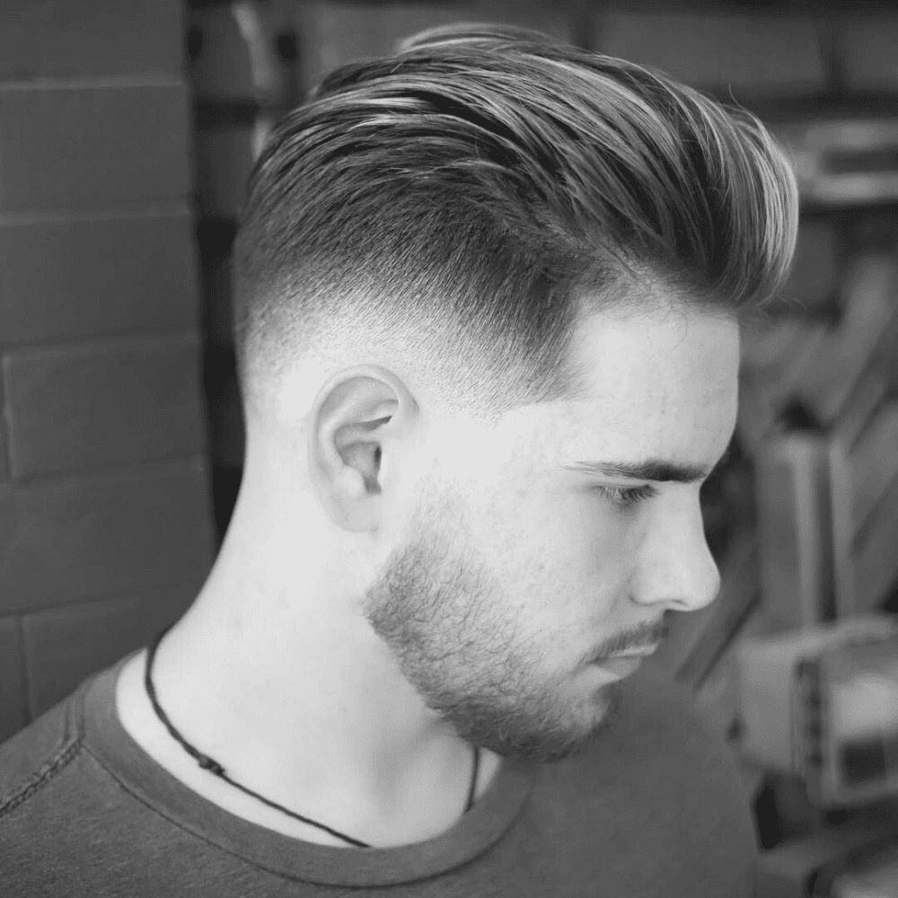 A three-quarter profile shot of a young man outdoors. He has a slick back haircut with blonde highlights and a mid-skin fade on the sides. He has a short beard and is wearing a purple t-shirt.
