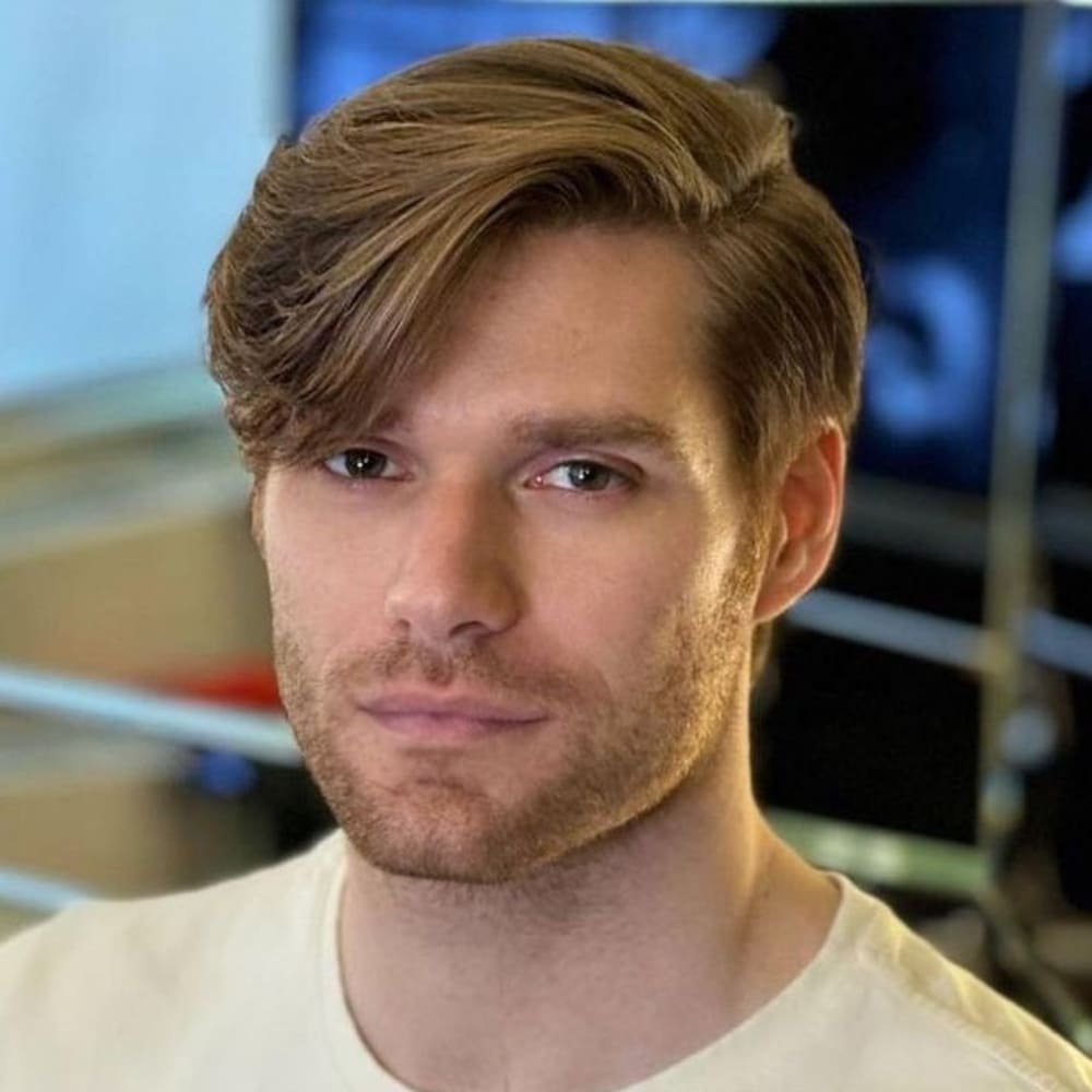 A man is depicted with a side-parted fringe hairstyle, featuring thick, light brown hair that is neatly swept to one side. The fringe is voluminous and flows naturally, framing the right side of his face, while the rest of the hair is smoothly styled to follow the side part. The hair on the sides is shorter and tapers slightly, creating a clean and polished look. His beard is closely trimmed, complementing the well-groomed appearance of his hairstyle. The man’s expression is relaxed and confident, with his soft eyes adding to the approachable and modern vibe of his look. He is dressed in a light-colored shirt, and the background is slightly blurred but suggests an indoor setting, possibly a contemporary or stylish space. The side-parted fringe adds a classic yet trendy element to his overall appearance, making it a versatile hairstyle suitable for various occasions.
