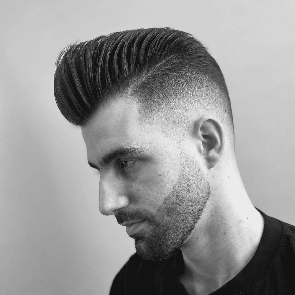 A black and white profile shot of a man with a dramatic slick back pompadour hairstyle. The hair has extreme volume at the front and is combed back with deep, visible lines. The sides feature a high skin fade that seamlessly blends into a sharply lined-up beard.