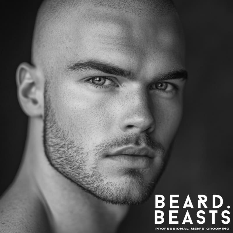 A black and white close-up portrait of a man with an oblong face shape, sporting a stubble beard that enhances his jawline while maintaining a natural and masculine appearance. The evenly trimmed light stubble adds subtle definition without elongating the face, making it one of the best beard styles for oblong faces. His sharp facial features and well-groomed beard create a balanced, structured look. He has a shaved head, which further accentuates the stubble beard’s ability to add texture and contrast. The Beard Beasts logo is displayed in the bottom right corner, reinforcing a premium men’s grooming aesthetic.