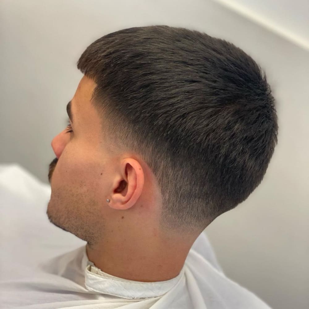 A close-up side profile of a man with a taper fade style. The style features a longer length on top, gradually tapering down to a clean fade around the sides and back of the head. The top is left with a smooth, even texture, while the fade blends seamlessly into the skin, creating a sleek and polished look. The hairline at the forehead is softly rounded, and the overall style is neat and precise. The man is wearing a white barber's cape, and the background is blurred, keeping the focus on the sharp details of the taper fade. The style is modern and stylish, suitable for a clean, professional appearance.