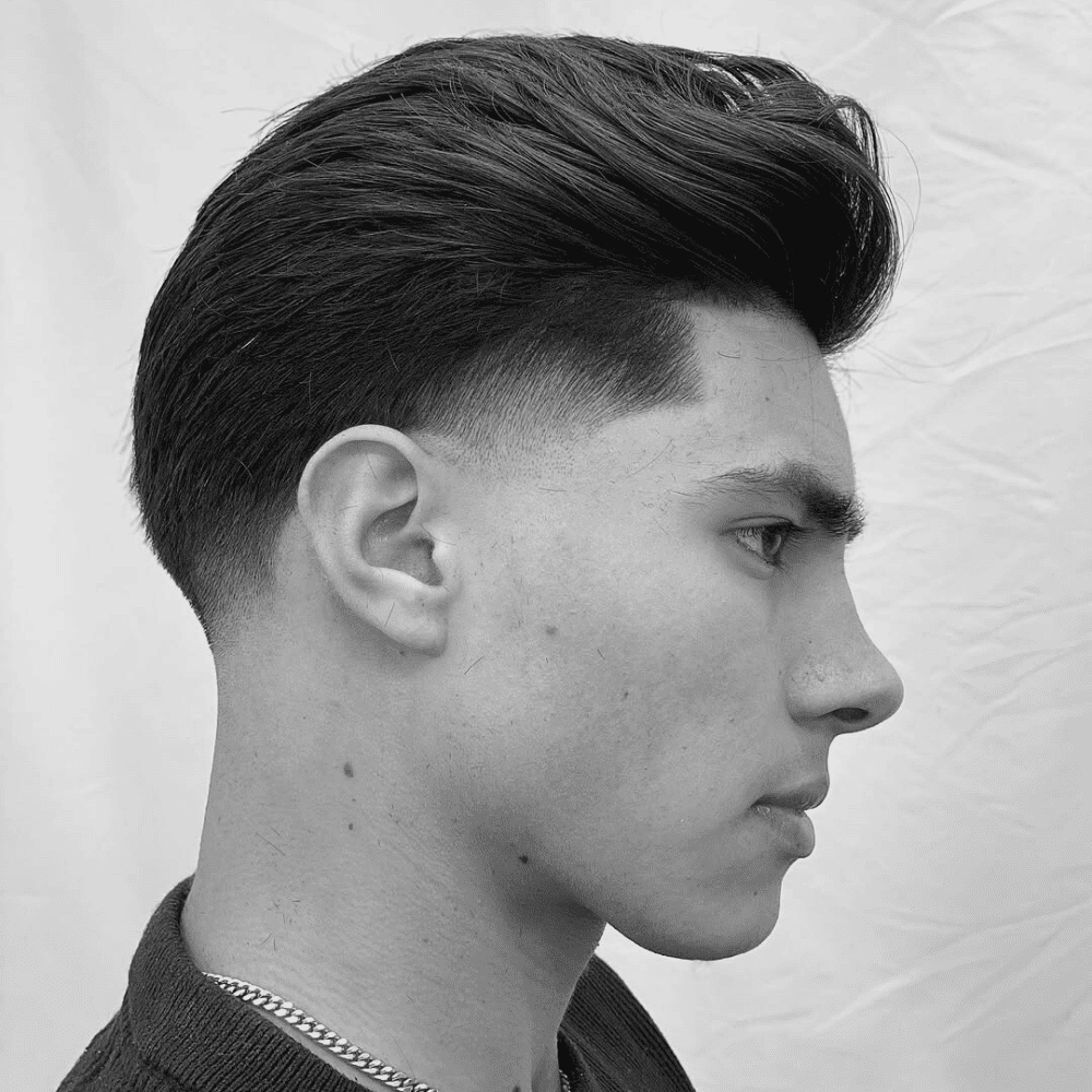 A black and white profile shot of a young man with a slick back taper fade haircut. The hair on top is voluminous and styled back, while the sides and back are neatly tapered short. The hairline is sharply defined.