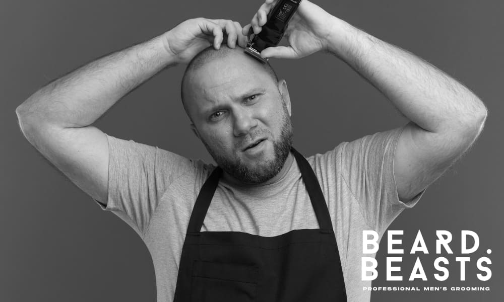 an using hair clippers to give himself a buzz cut at home, wearing a black apron and gray t-shirt – Beard Beasts professional men’s grooming