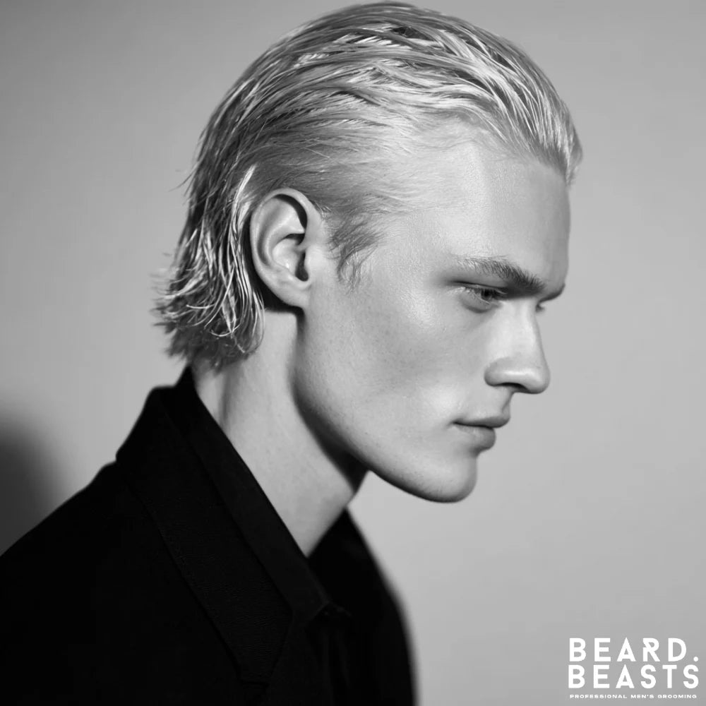 A studio portrait of a man with platinum blonde hair styled in a wet-look slick back. He is shown in three-quarter profile, looking down, and is wearing a black collared shirt.