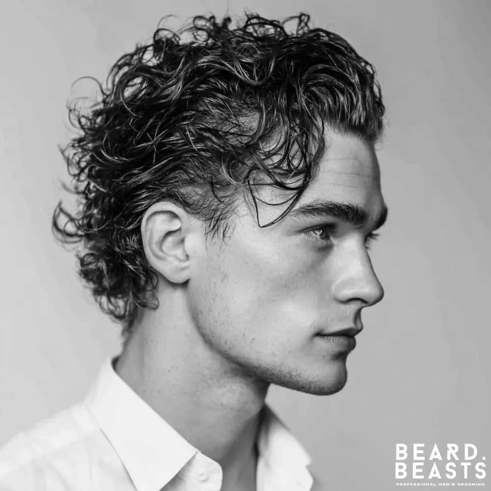 A close-up studio portrait of a man in profile. He has medium-length, curly brown hair with a wet look, styled back from his face while allowing some curls to frame his forehead. He has blue eyes, light stubble, and is wearing a white collared shirt.