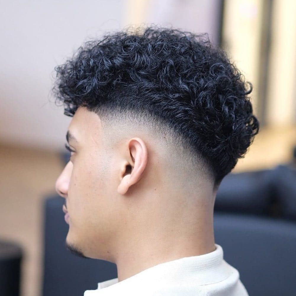 A close-up image of a young man with neatly trimmed facial hair, featuring a stylish haircut that seamlessly transitions from shorter hair on the sides to fuller, well-defined curls on top.