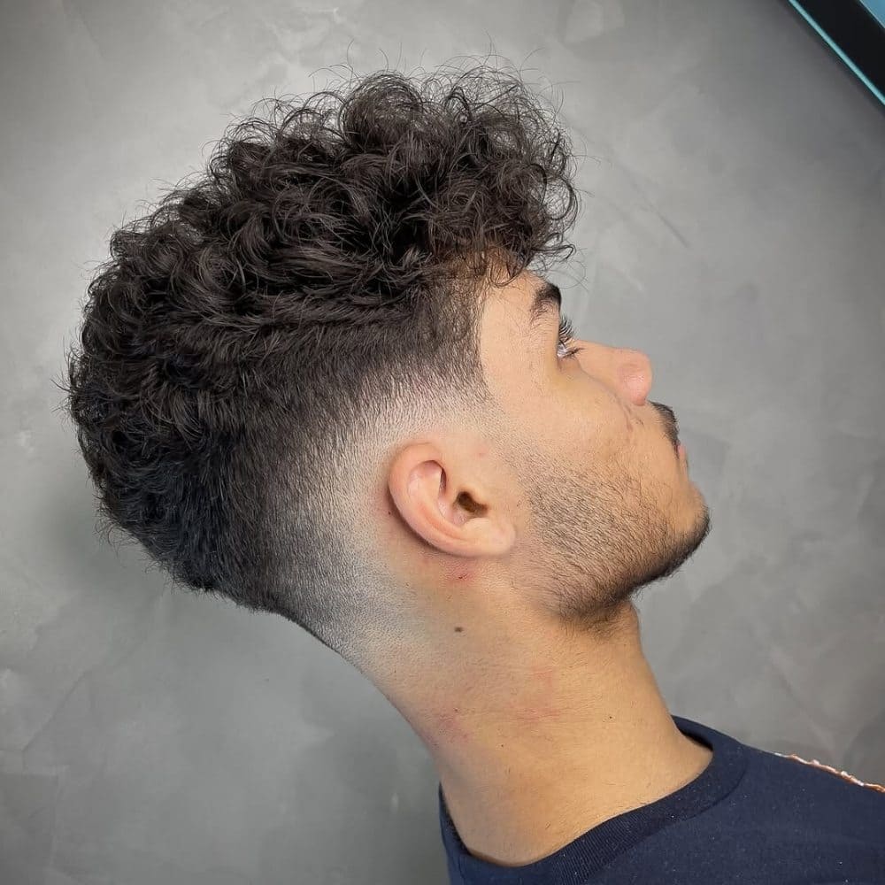 This image features a stylish curly low fade haircut, where the hair transitions smoothly from fuller curls on top to a closely cropped, tapered fade at the sides and back. The fade starts just above the ears, blending seamlessly into the natural curls, creating a clean and modern look. This haircut is perfect for highlighting the texture of curly hair while maintaining a sleek, low-maintenance style. Ideal for those seeking a balance between a sharp fade and defined curls.