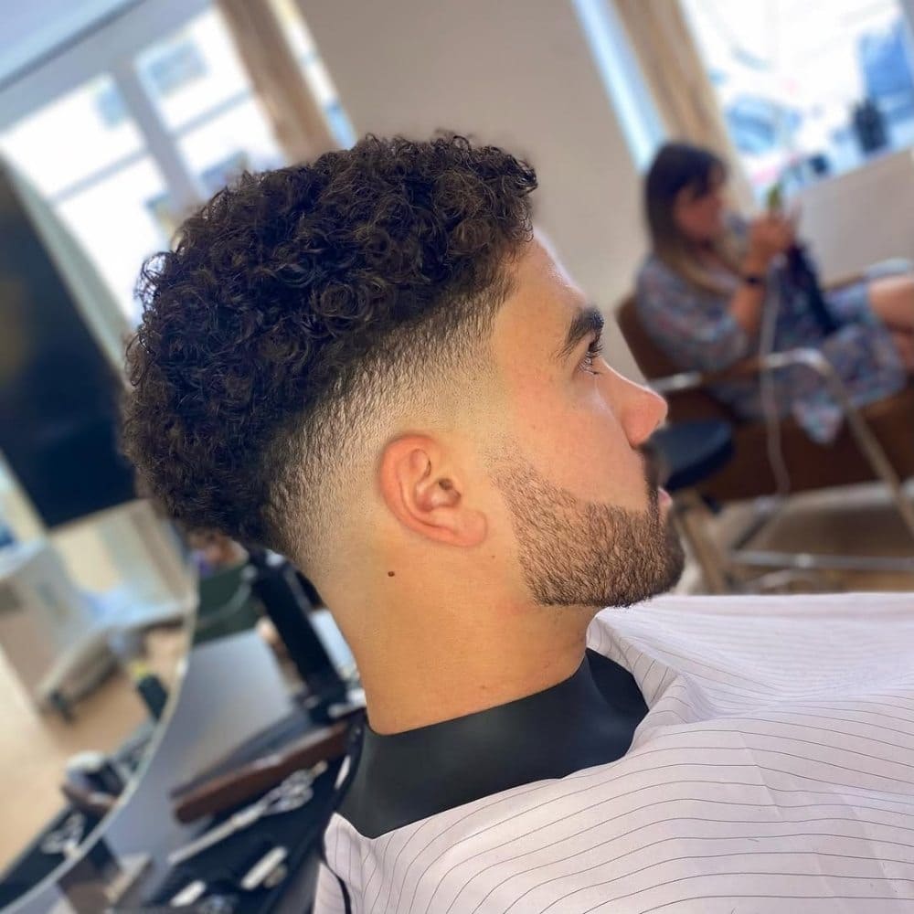 Curly mid fade hairstyle featuring a smooth transition from shorter faded sides to voluminous curls on top. This stylish look, ideal for men with curly hair, provides a sharp, modern appearance while maintaining the natural texture of the curls. The fade starts around the mid-point of the head, accentuating the curls on top. Perfect for those seeking a fresh and trendy curly hair fade.