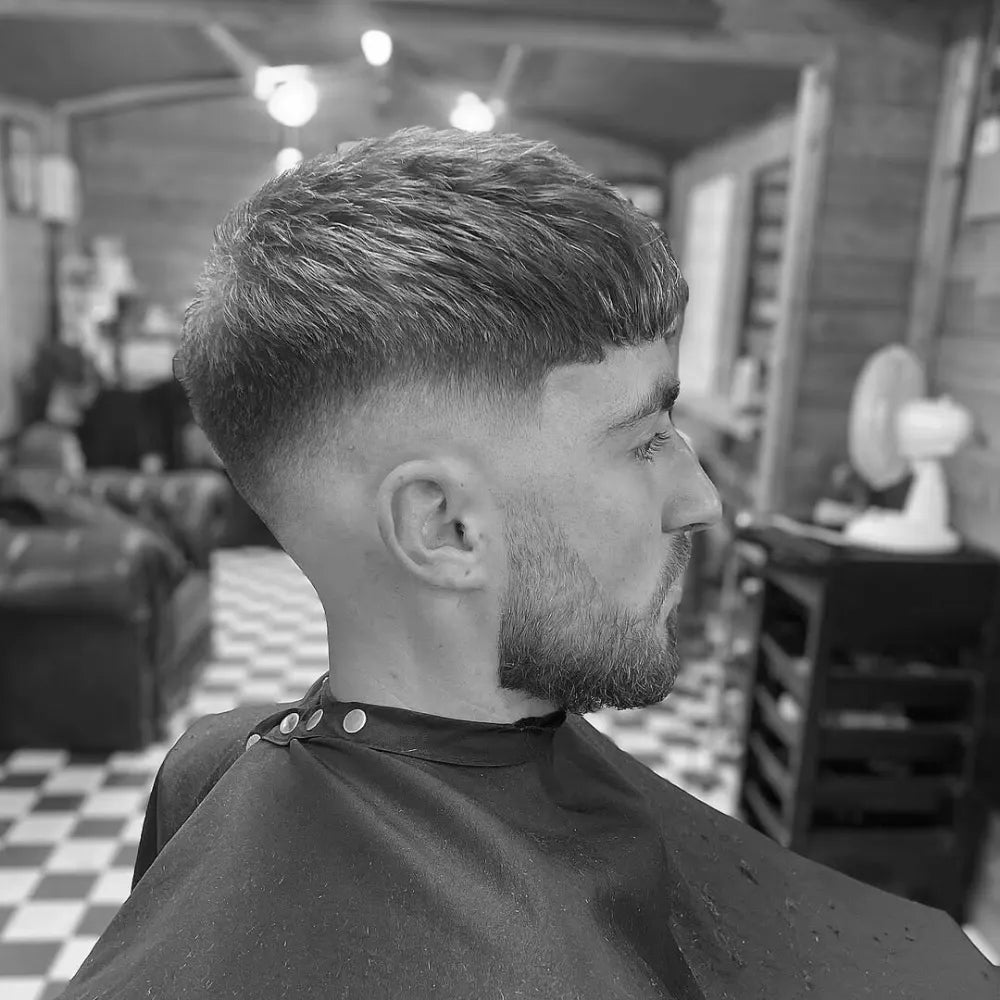 Man with a mid drop fade and long textured crop, featuring a sharp fade that curves behind the ears and blends into a neatly faded beard for a modern, stylish look.