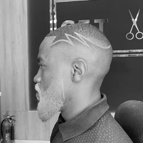 A bold high fade haircut with a unique hair design, featuring sharp, intricate razor-cut patterns on the side for a stylish and artistic touch. The crisp fade seamlessly blends into the skin, complementing the well-groomed, full beard. This modern fade haircut is perfect for men looking to showcase creativity and precision in their hairstyle.