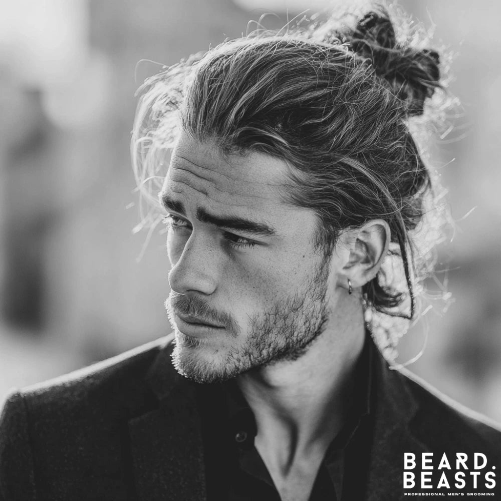 Man with layered long hair styled into a man bun, wearing a dark jacket, with soft flyaways framing the face.