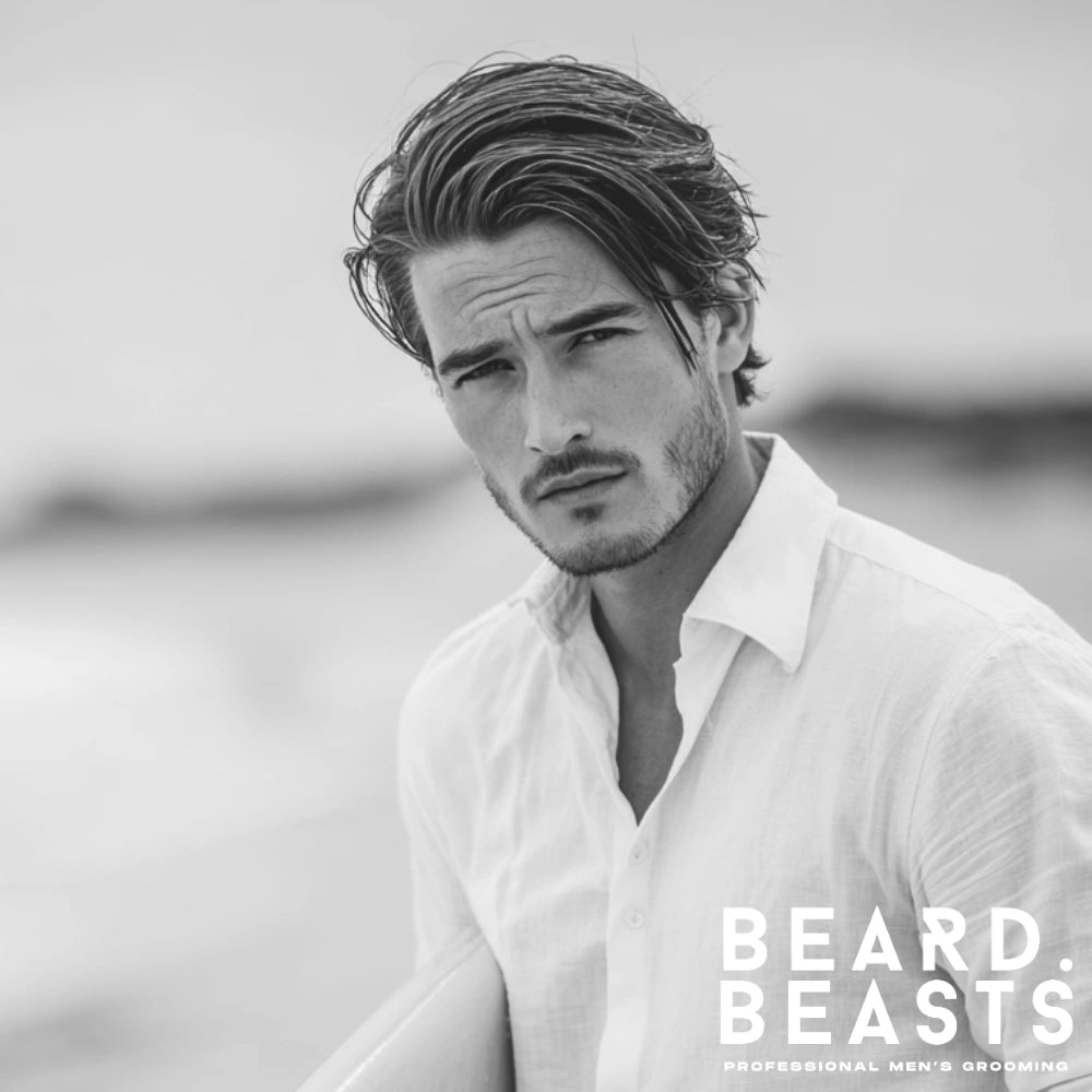 Man with long comb over surfer hairstyle holding a surfboard on the beach.
