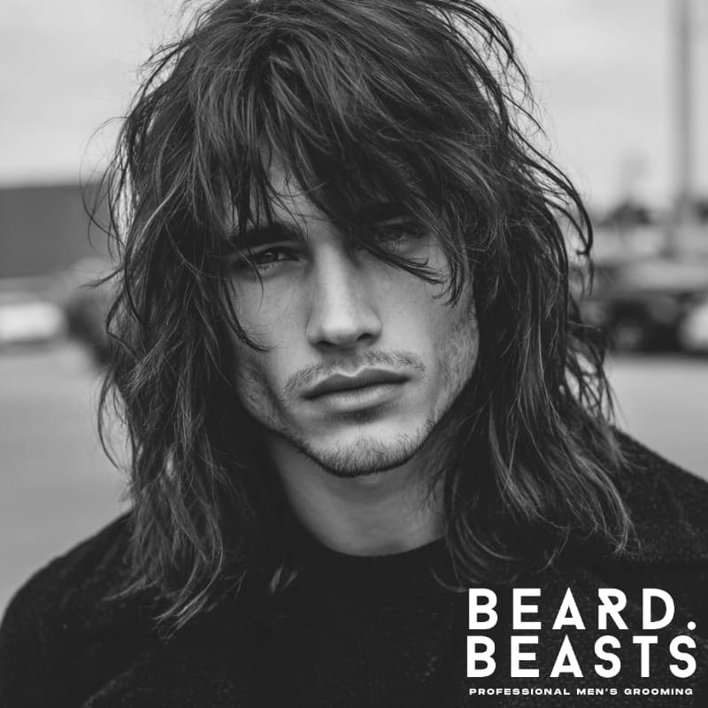 A modern men’s hairstyle featuring long, layered, and textured hair with bangs falling naturally over the forehead. Perfect for those seeking a relaxed, tousled look with volume and movement, ideal for adding depth and style to long hair.