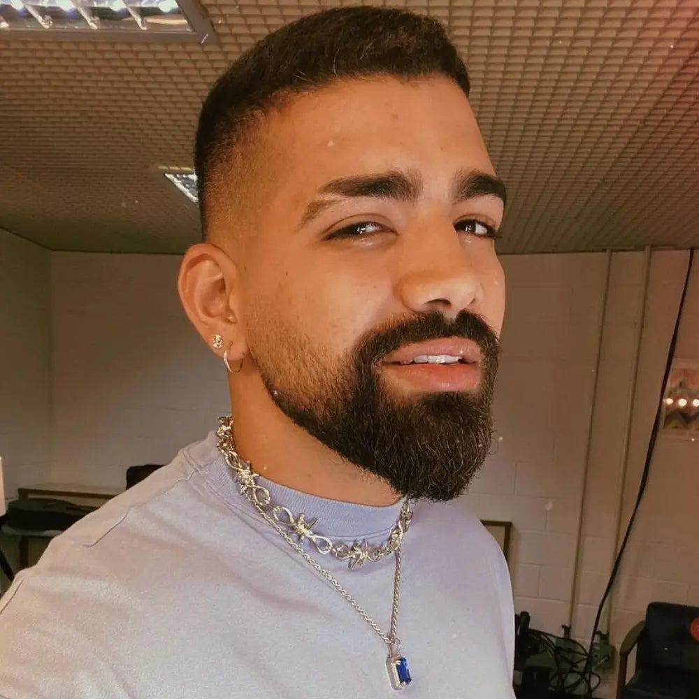 Man with an extended goatee and faded sides, featuring a well-groomed beard and clean neckline, wearing layered necklaces.