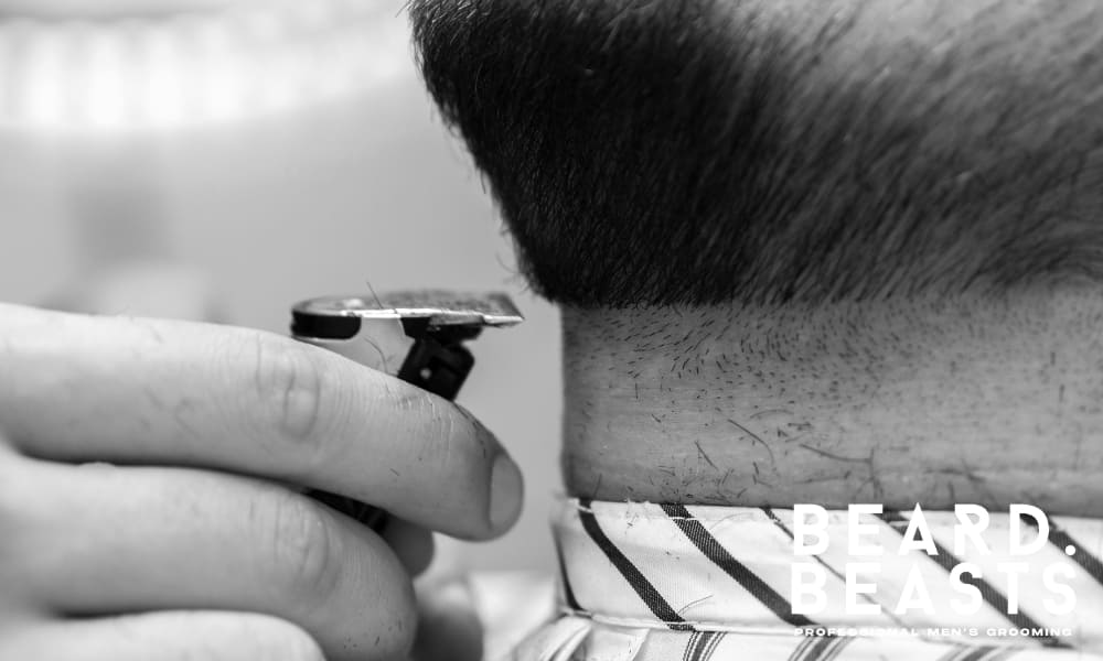 A black and white close-up image of a barber precisely trimming a man's beard neckline with a straight razor, ensuring a clean and well-defined shape. The sharp, even neckline creates a polished and structured look, which is essential for maintaining a well-groomed beard style. The contrast between the trimmed and untrimmed areas highlights the importance of neckline maintenance in professional beard grooming.