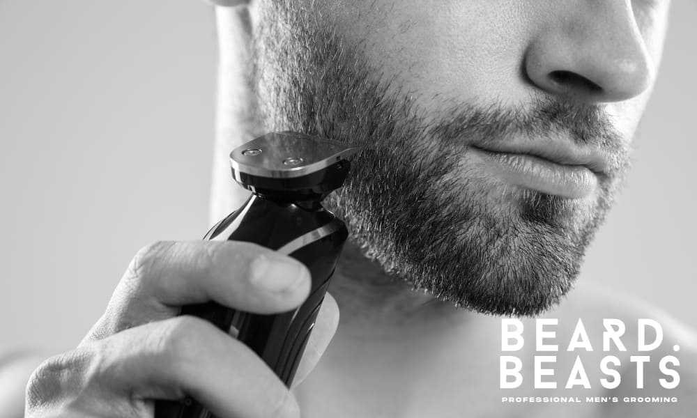 A black and white close-up of a man carefully trimming his beard with an electric trimmer, focusing on shaping and maintaining a clean, well-groomed look. His evenly maintained beard enhances his oblong face shape, ensuring balance and structure without adding unnecessary length. The precise trimming technique highlights the importance of regular grooming for achieving a sharp and masculine appearance.