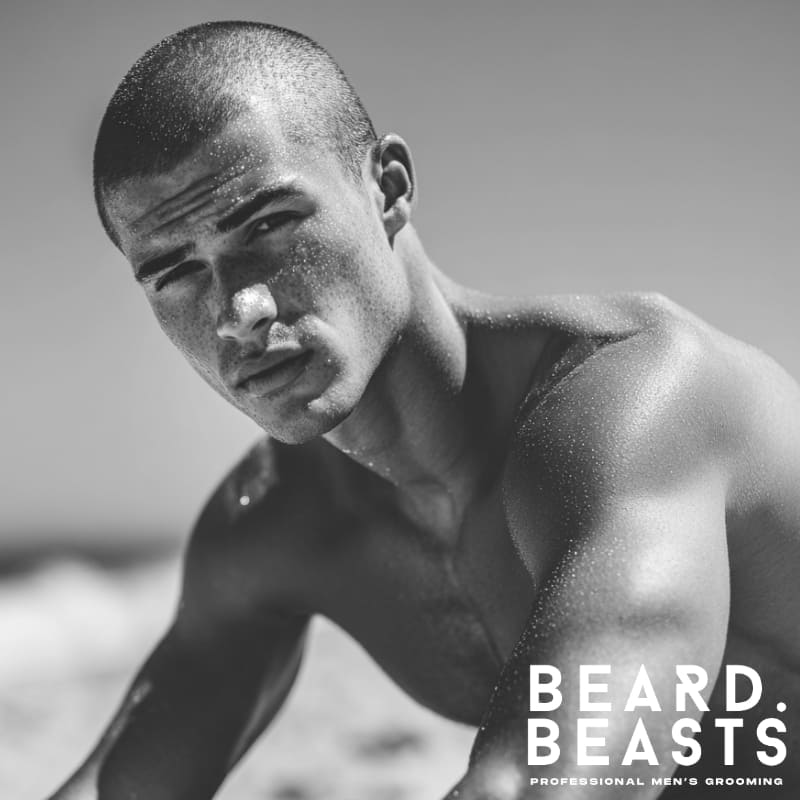 Portrait of a male model with a very short buzz cut, with subtle texture from water droplets, looking directly at the viewer on a beach.