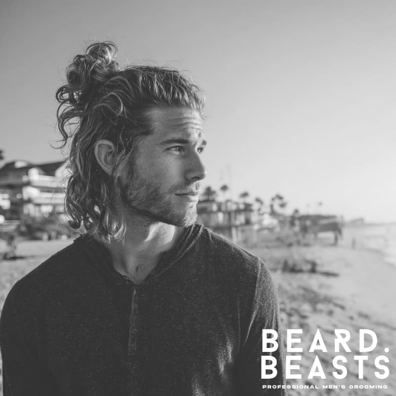 Male model with long, wavy blonde hair styled in a half-up, half-down surfer look, standing on a sunny beach.