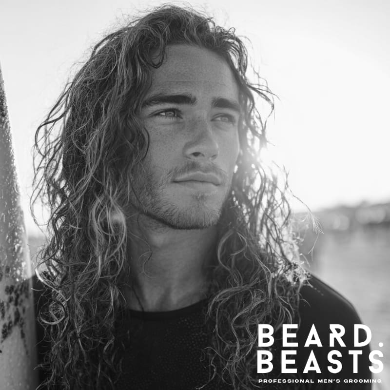 Portrait of a male model with long, voluminous, textured curly hair, backlit by the setting sun on a beach, embodying a natural surfer look.