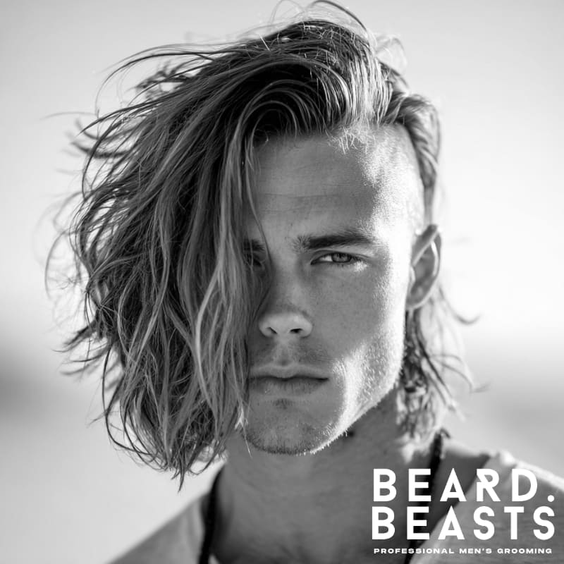 Close-up of a male model with long, wavy blonde hair flowing to one side, showcasing a distinct undercut on the other side, for a modern surfer hairstyle.