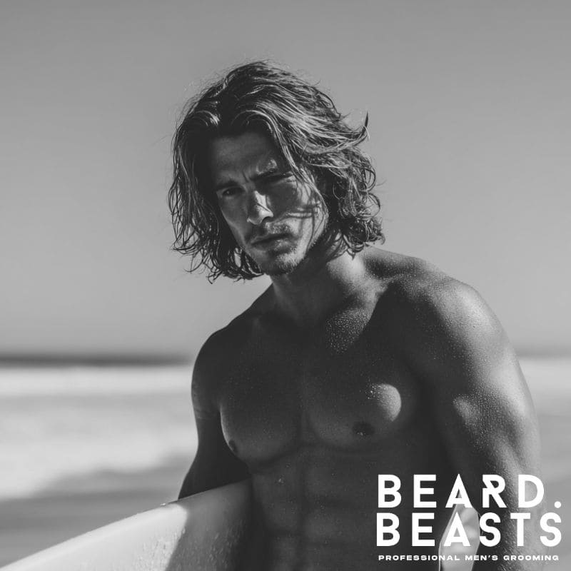 Male surfer with long, dark hair styled with a distinct middle part, standing on a beach with a surfboard, embodying a classic surfer aesthetic.